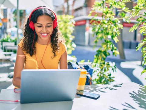 A student sits outside wearing headphones, smiling while working on a laptop with a drink and phone nearby.