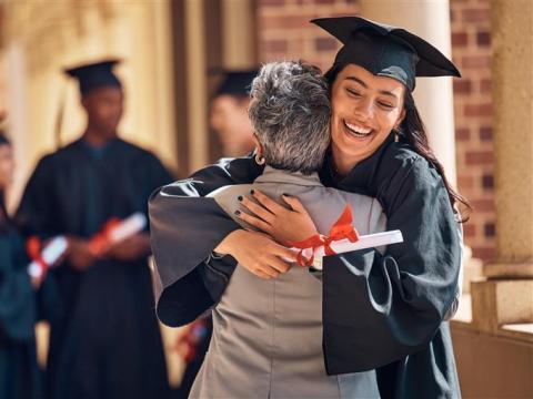 Graduate in cap and gown hugs an older woman while holding a diploma with a red ribbon. Other graduates celebrate in the background.