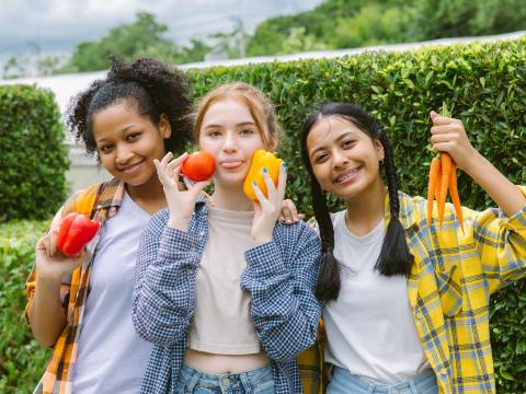 Three students hold vegetables in a garden.